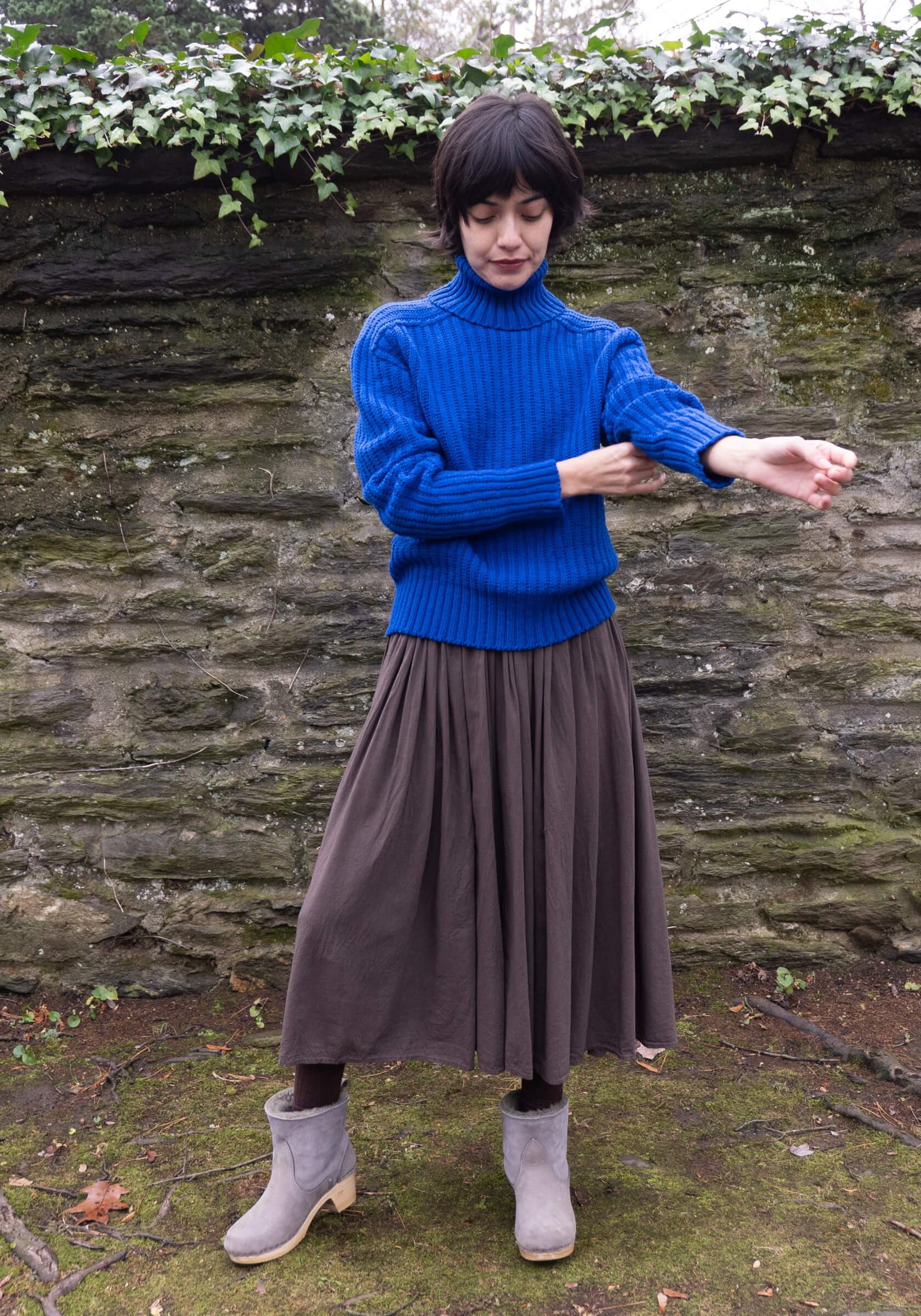 Person wearing a blue sweater and brown skirt standing in front of a stone wall with greenery.