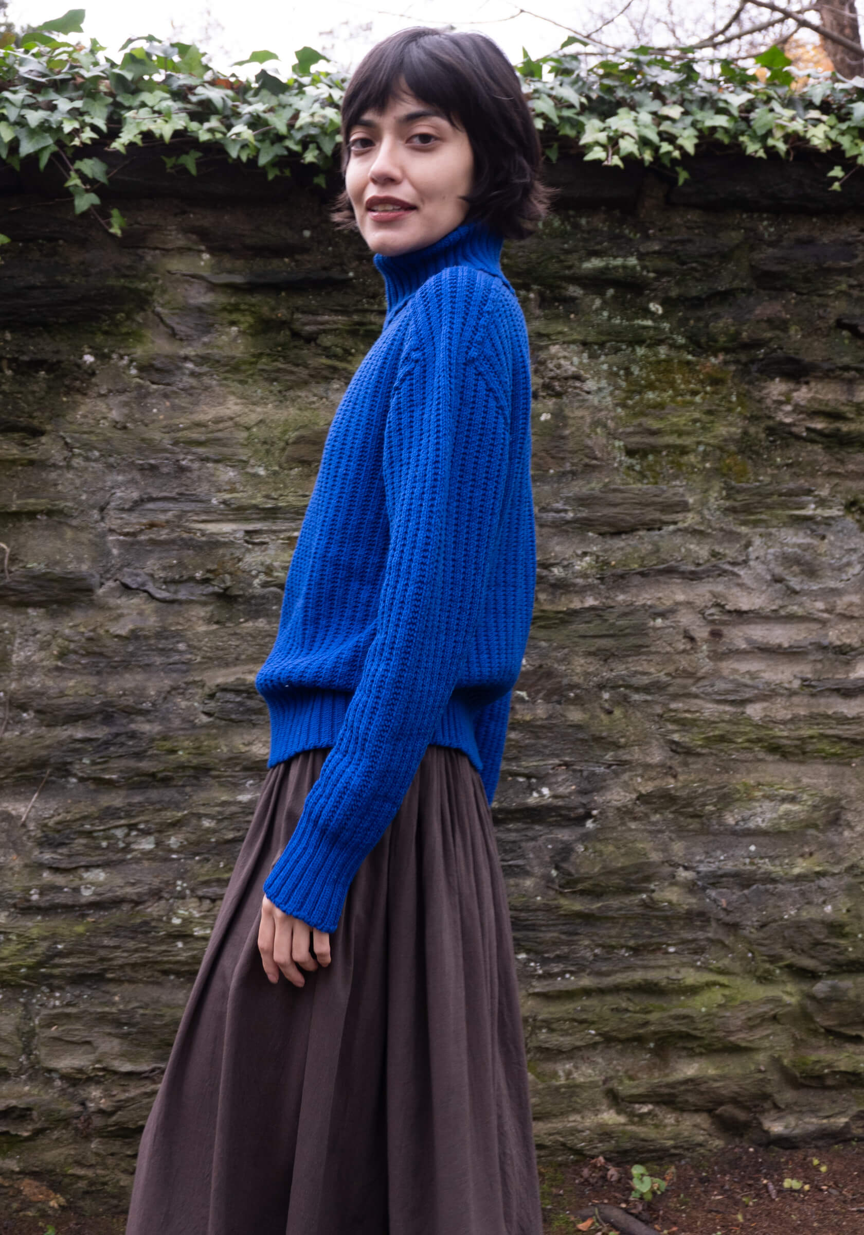 Person wearing a blue sweater and brown skirt standing against a stone wall with greenery.