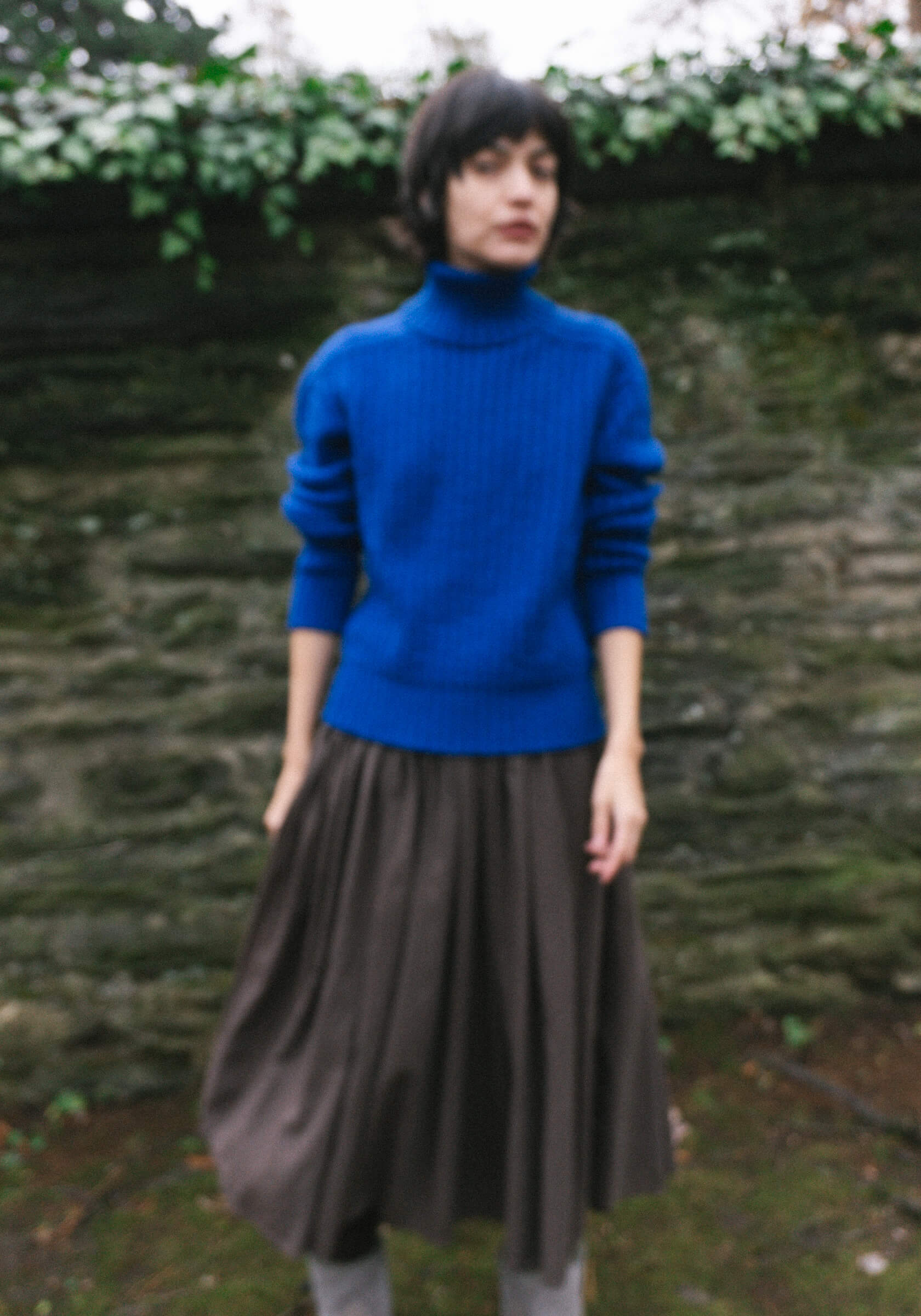 Person wearing a blue sweater and brown skirt standing in front of a stone wall with greenery.
