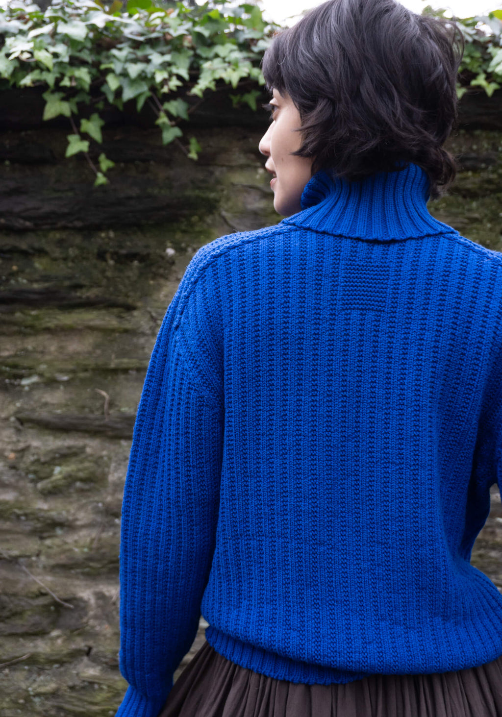 Person wearing a blue knitted sweater standing in front of a stone wall with greenery.