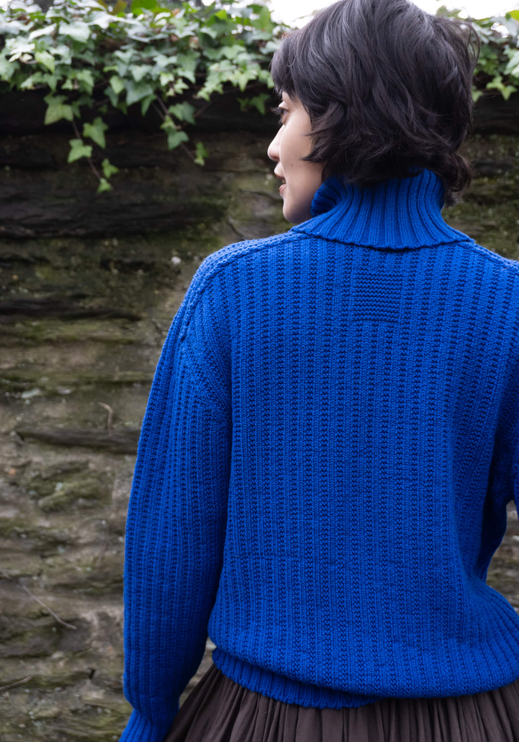 Person wearing a blue knitted sweater standing in front of a stone wall with greenery.