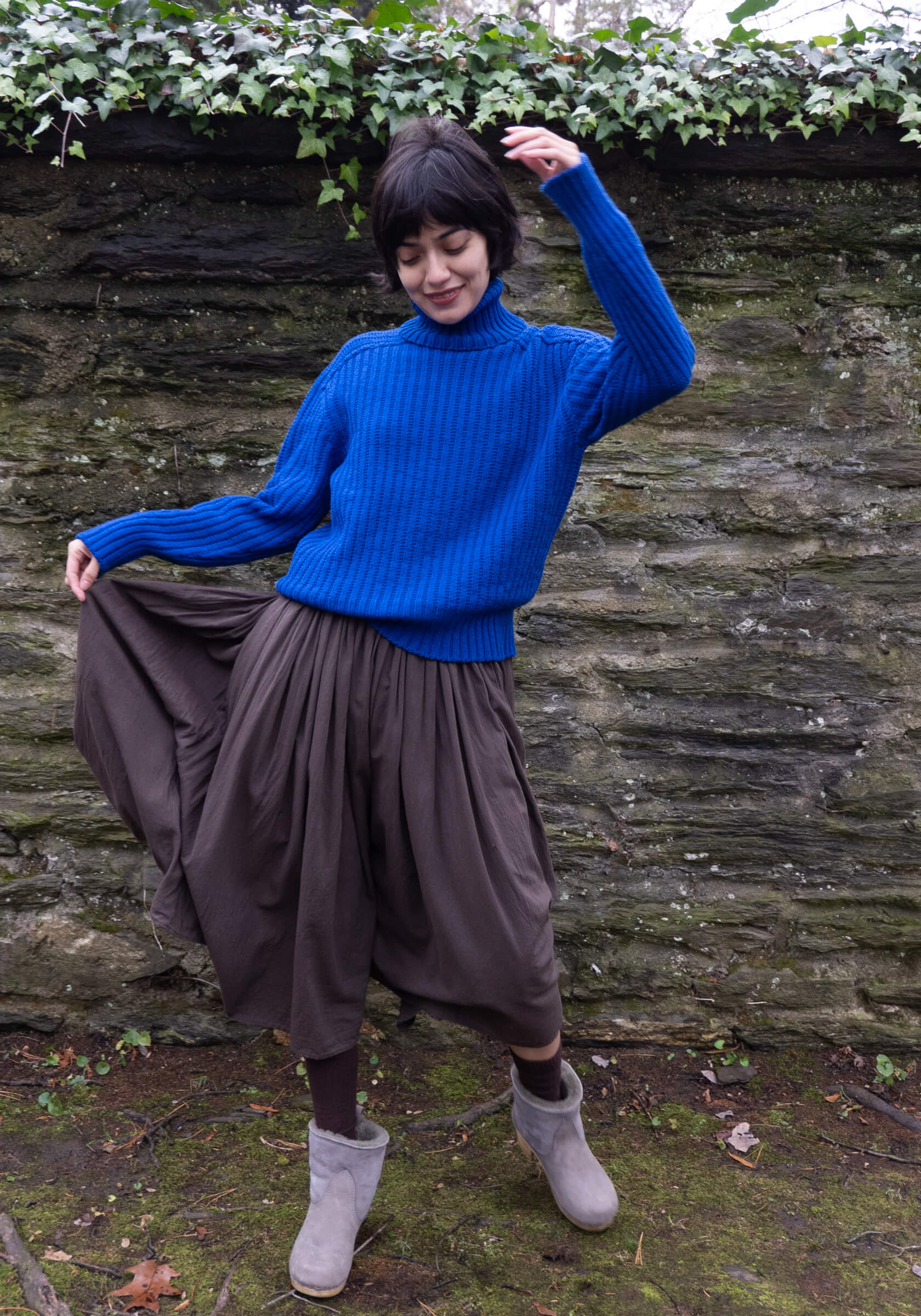 Person wearing a blue sweater and brown skirt standing against a stone wall with greenery.
