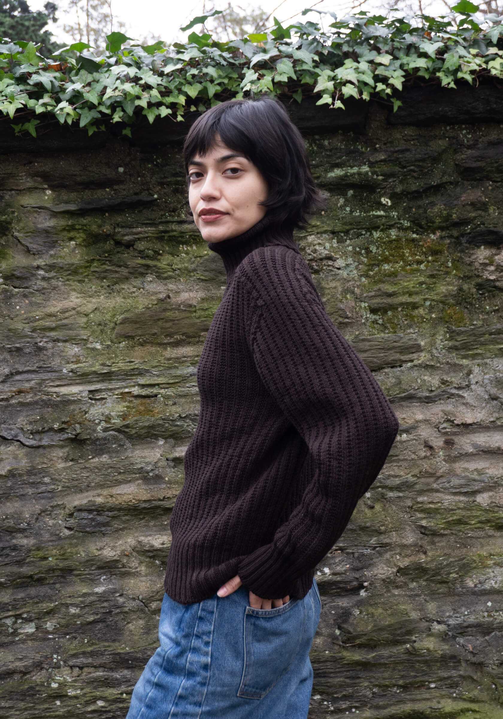 Person wearing a dark brown sweater and blue jeans standing against a stone wall with greenery.