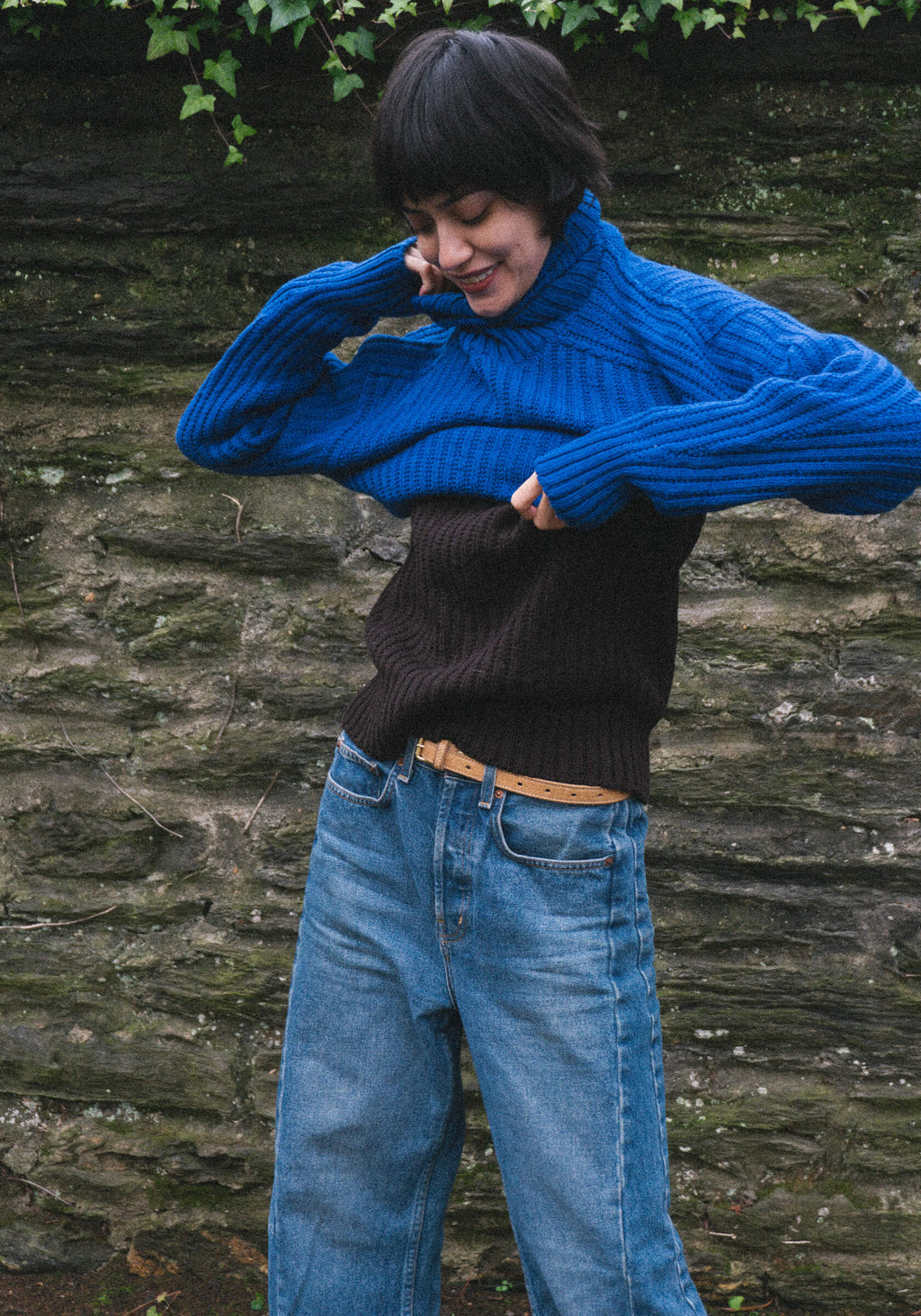 Person wearing a blue sweater over a black one, standing against a stone wall.