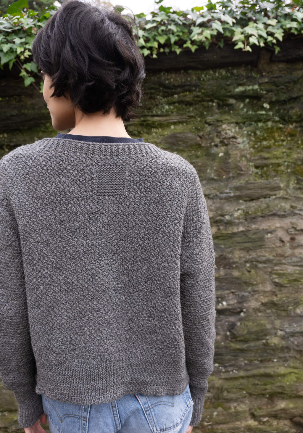 Person wearing a gray knitted sweater standing against a stone wall with greenery.