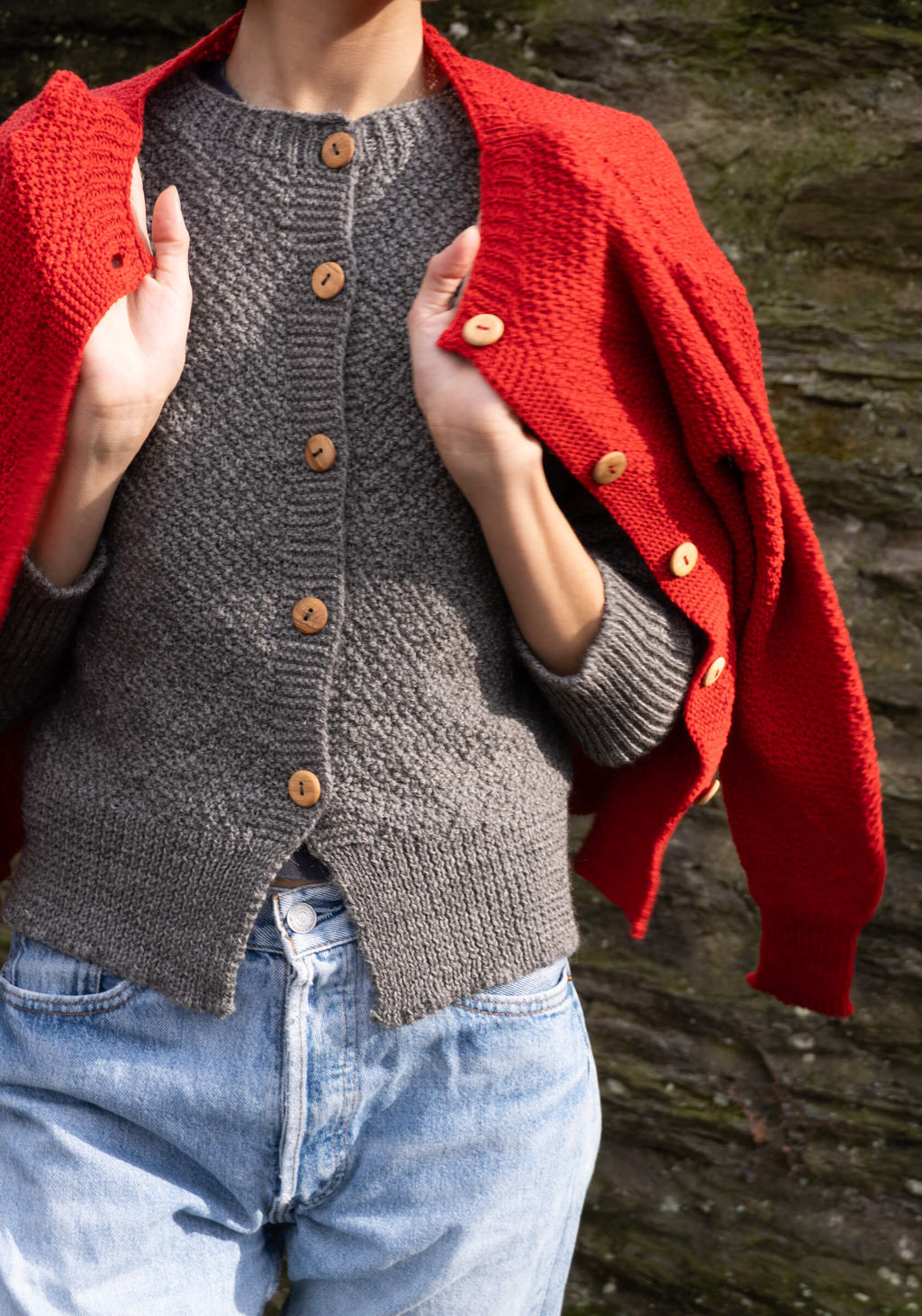 Person wearing a red cardigan over a gray knitted cardigan with wooden buttons, standing against a natural background.