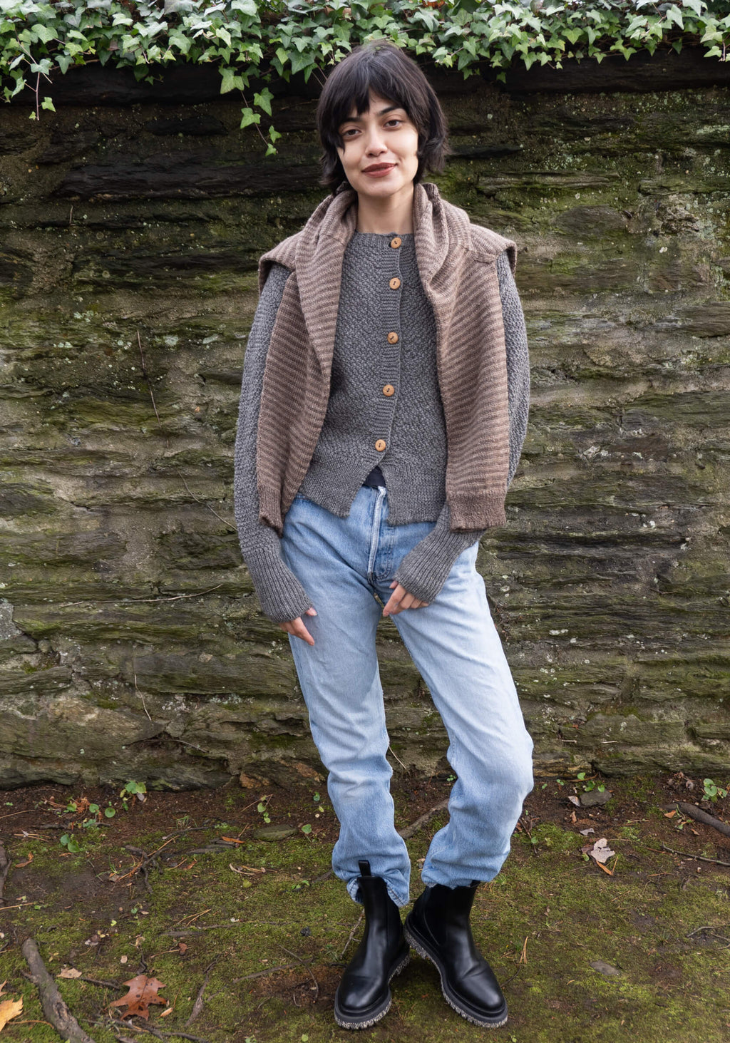 Person wearing a gray sweater, brown cardigan, and blue jeans standing against a stone wall.