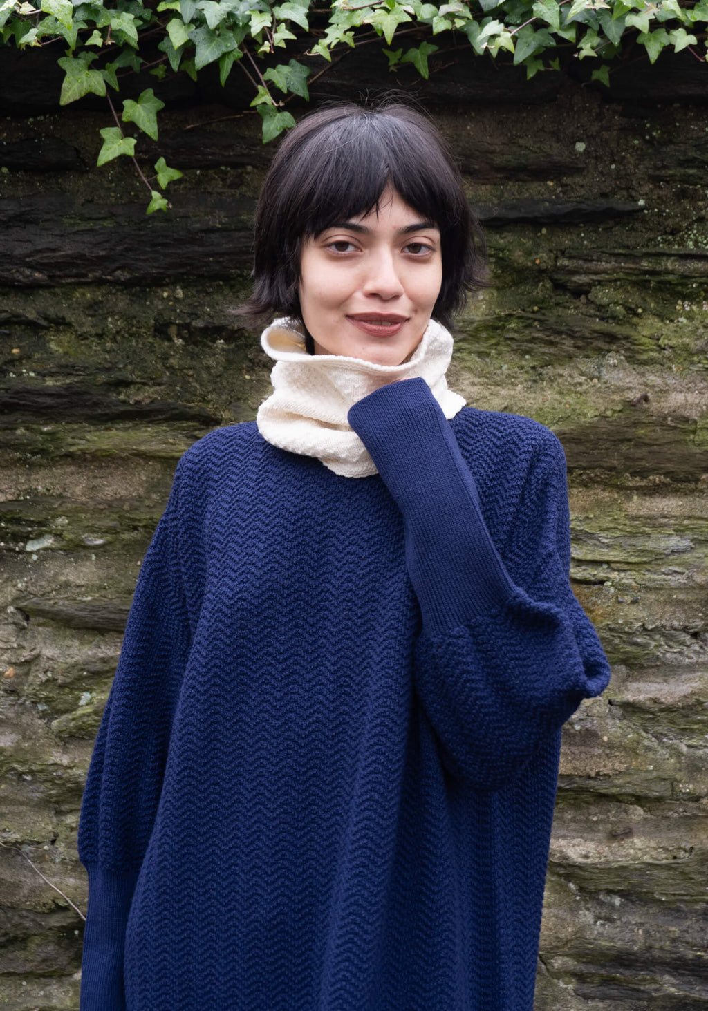 Woman wearing a blue sweater and white cowl neck scarf standing against a stone wall with greenery.