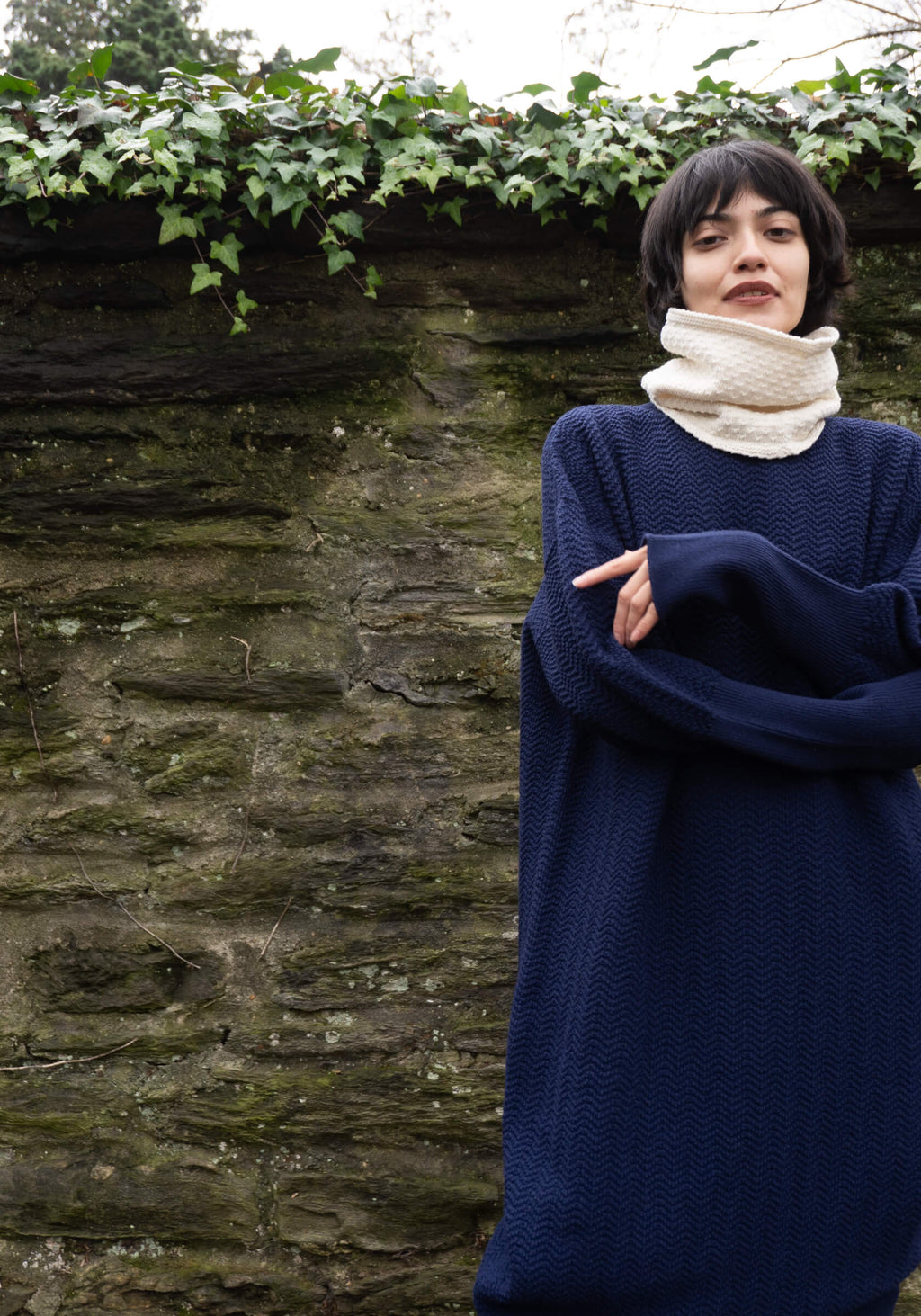 Person wearing a navy blue sweater and beige scarf standing against a stone wall with greenery above.