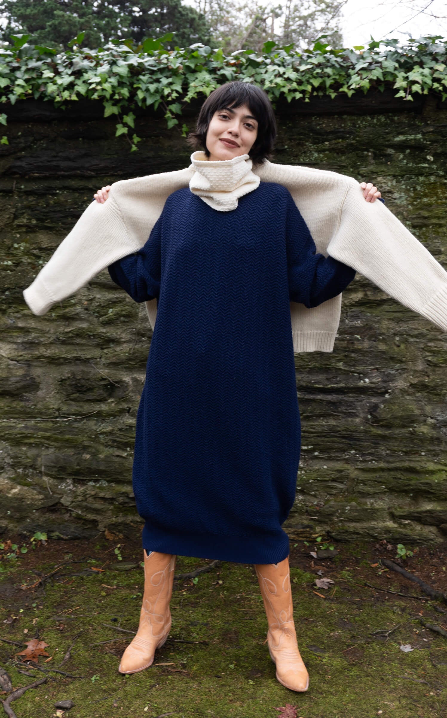 Person wearing a navy dress with a white sweater over shoulders against a stone wall background