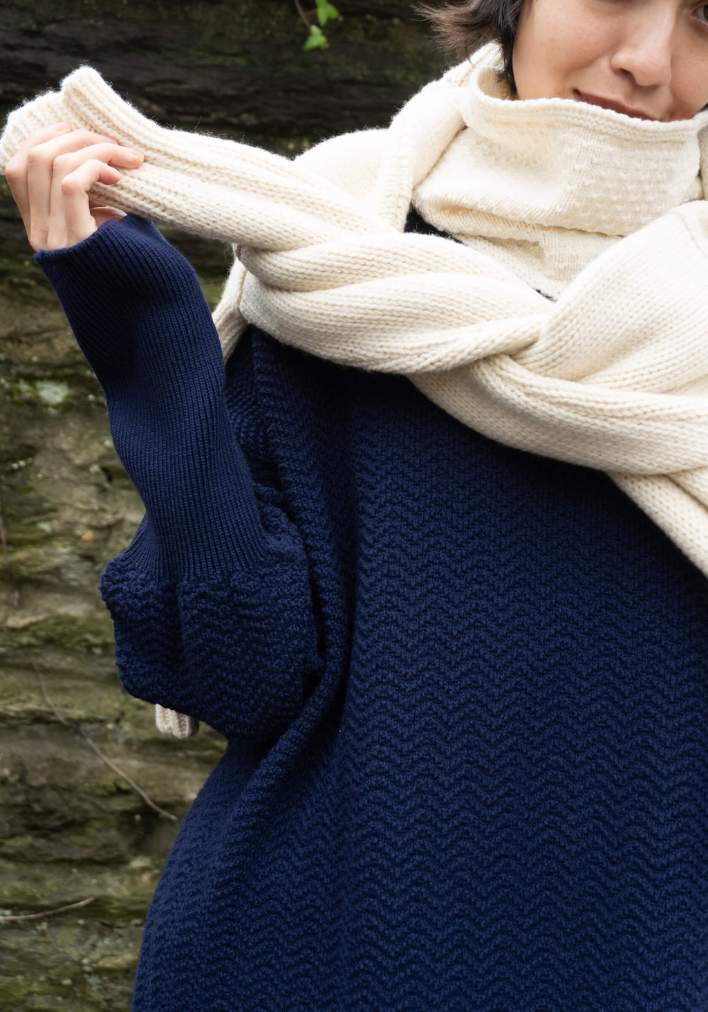 Person wearing a navy blue sweater and cream scarf against a stone wall background