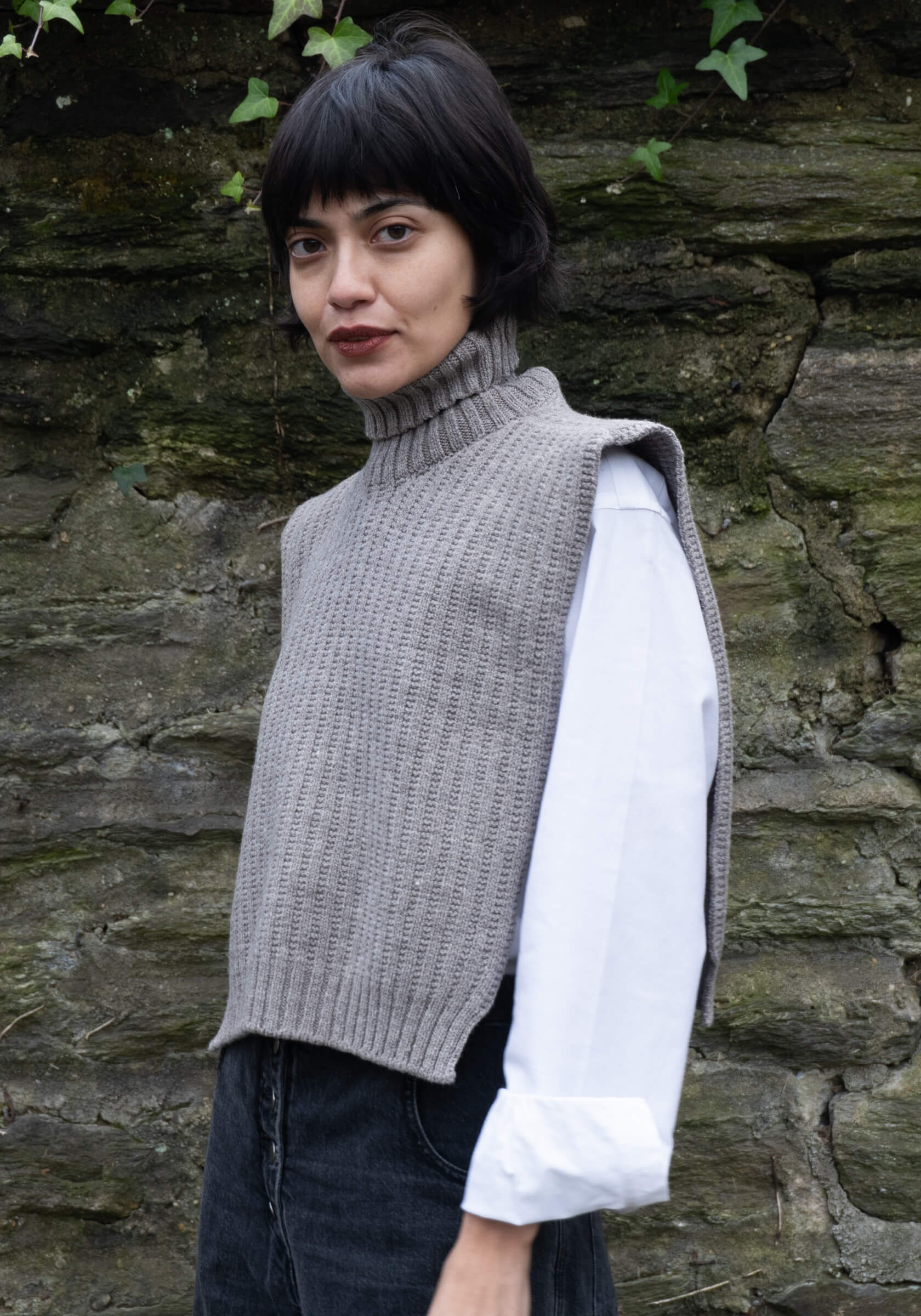 Woman wearing a gray knitted vest over a white shirt against a stone wall.