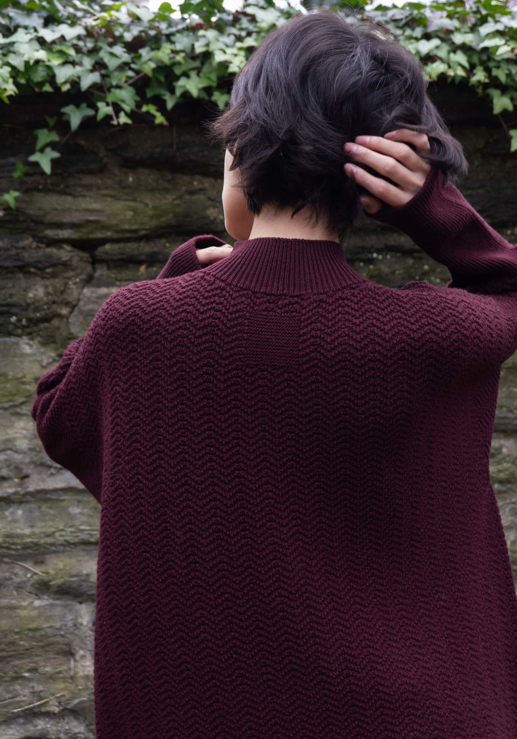 Person wearing a burgundy sweater against a stone wall with greenery.
