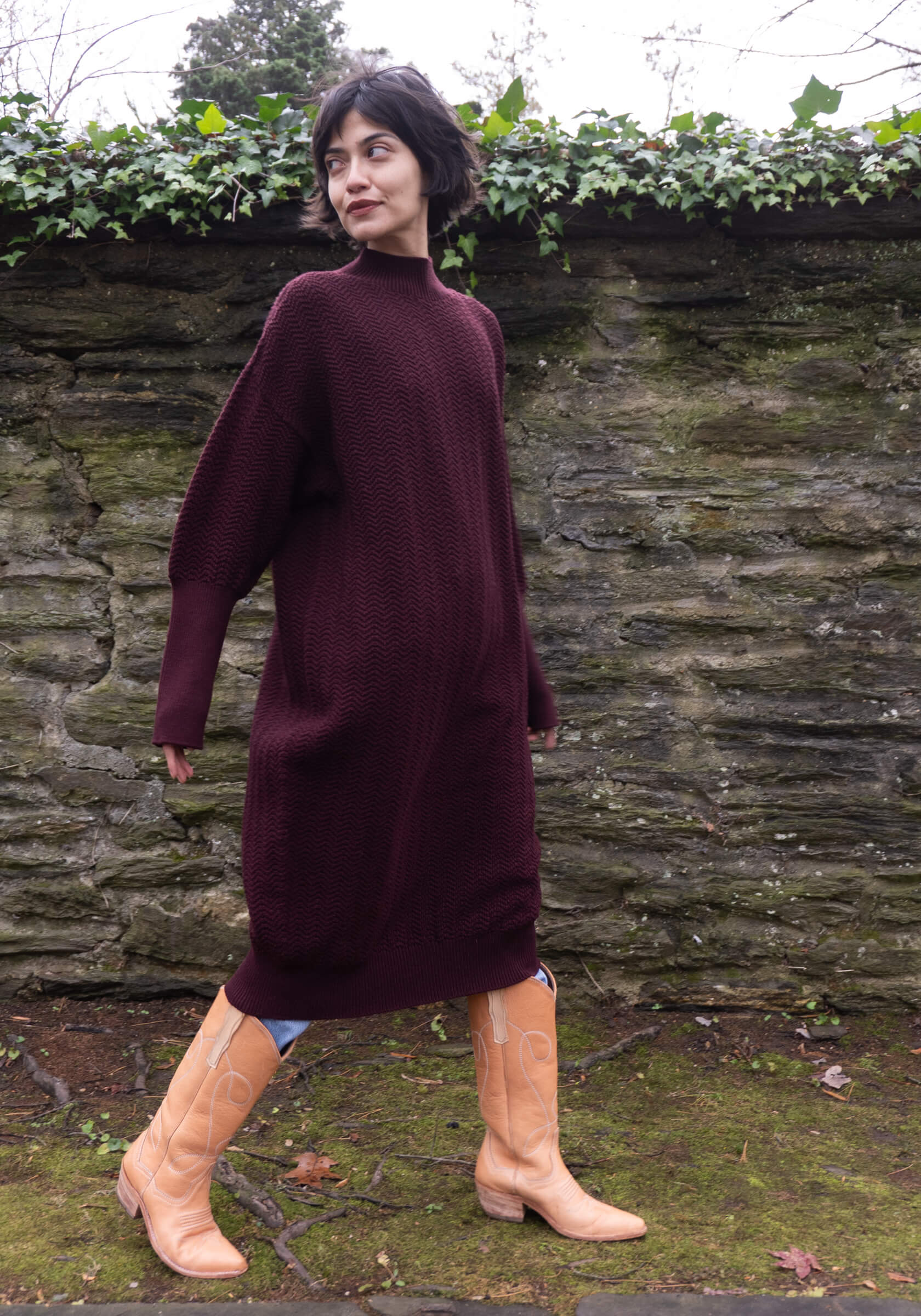 Woman wearing a long burgundy dress with beige boots standing against a stone wall.