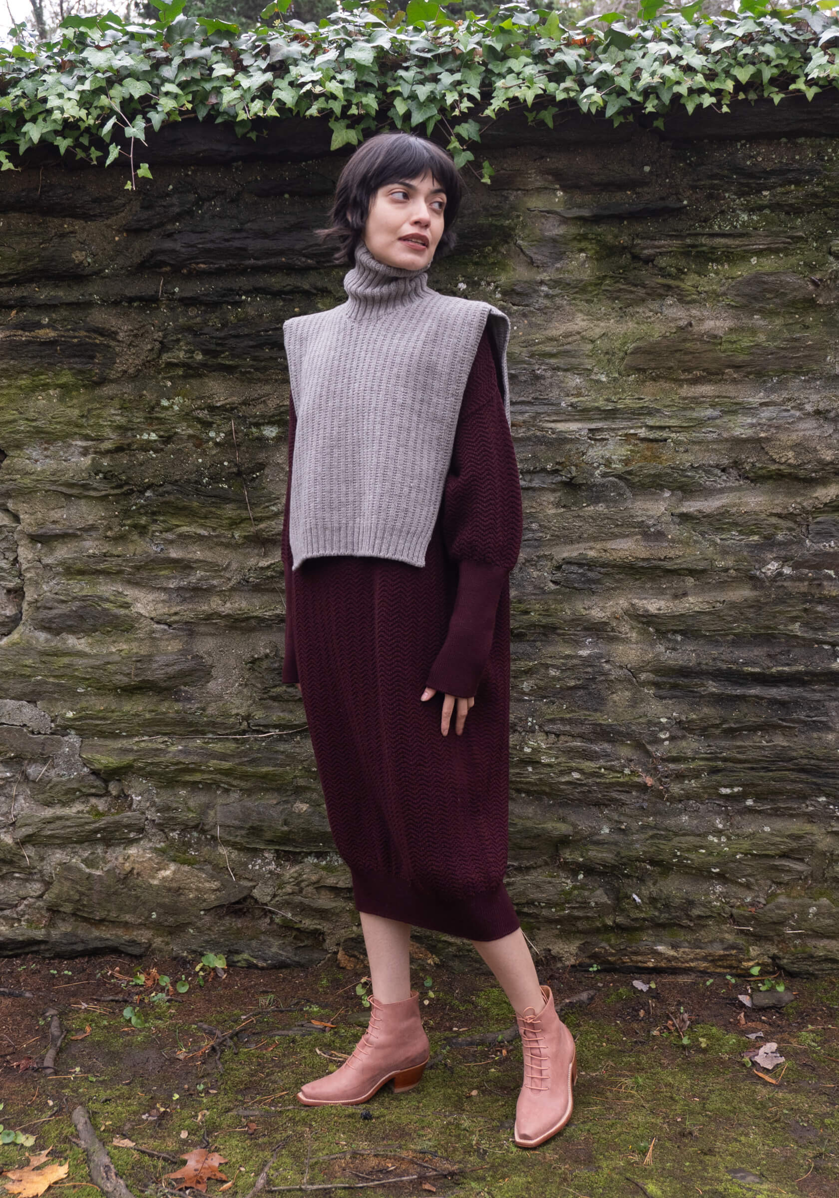 Person wearing a gray sleeveless sweater over a burgundy dress against a stone wall.