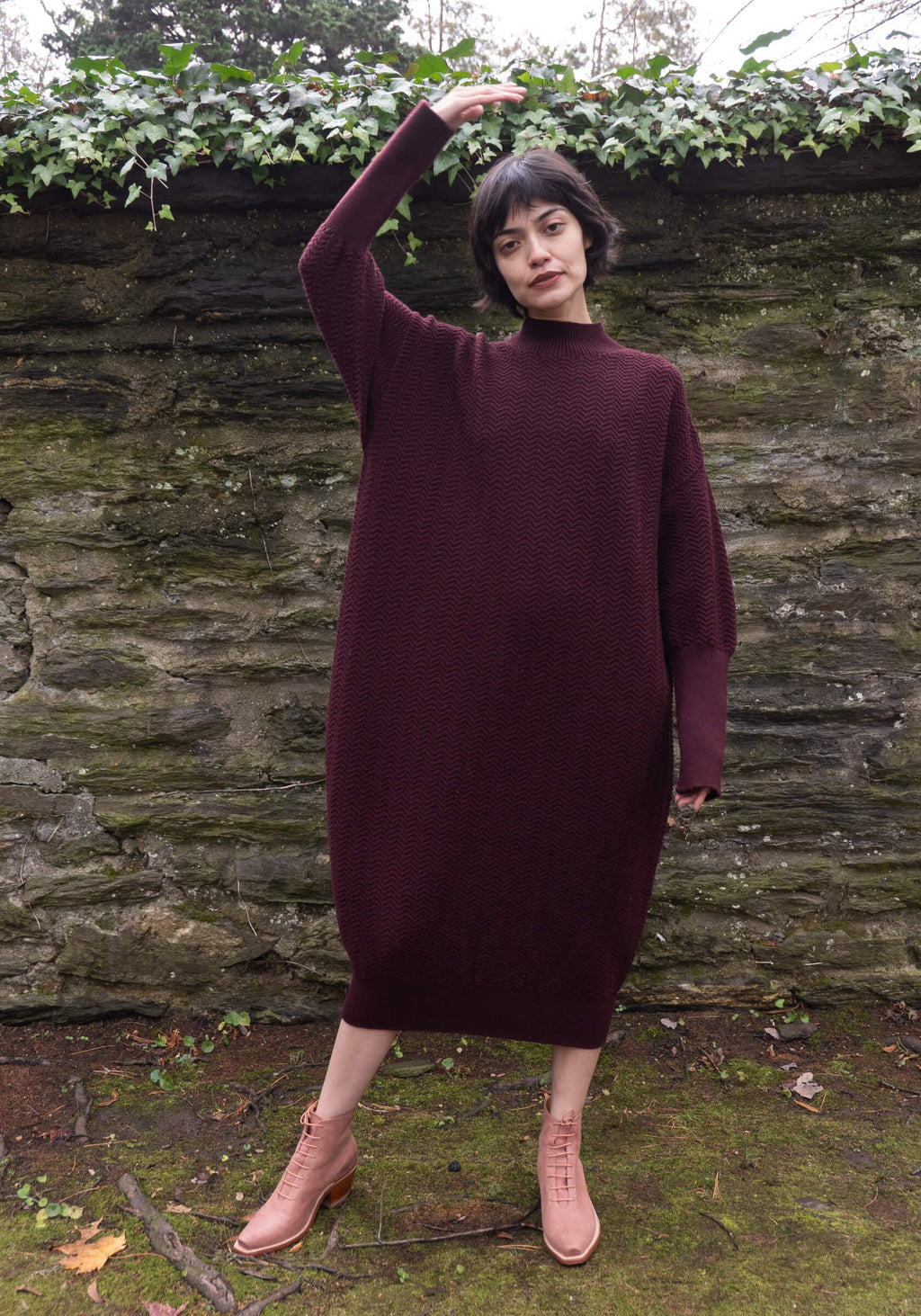 Person wearing a burgundy sweater dress standing in front of a stone wall with greenery.