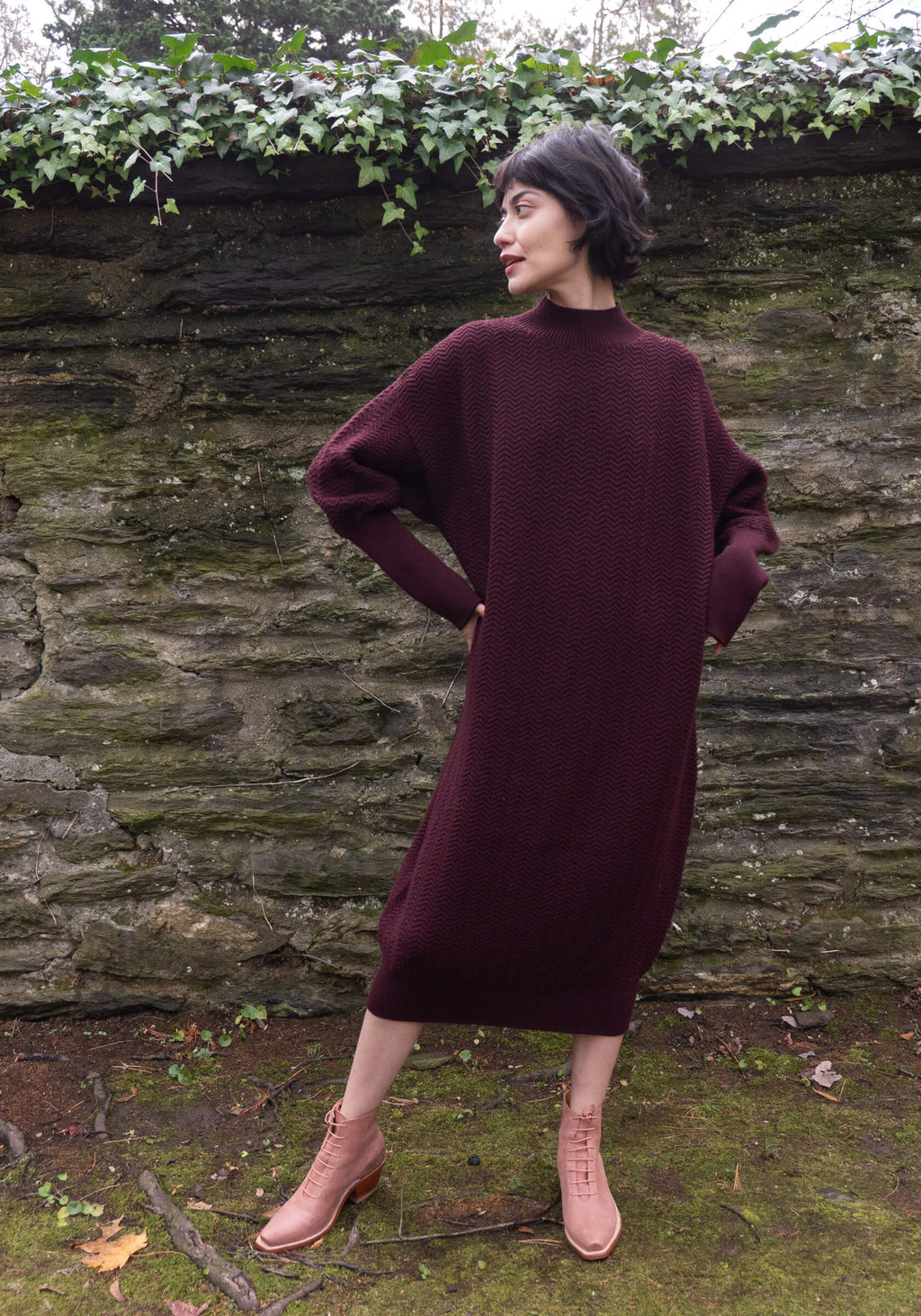 Woman wearing a burgundy sweater dress standing against a stone wall with greenery.