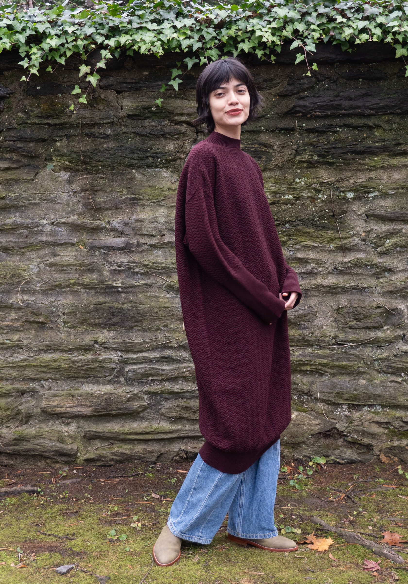 Person wearing a long burgundy sweater and blue pants standing against a stone wall with greenery.
