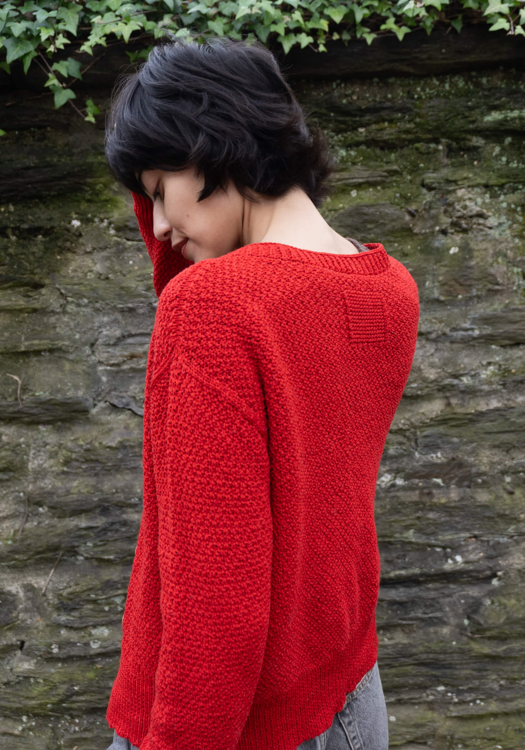 Person wearing a red sweater standing against a stone wall with greenery.