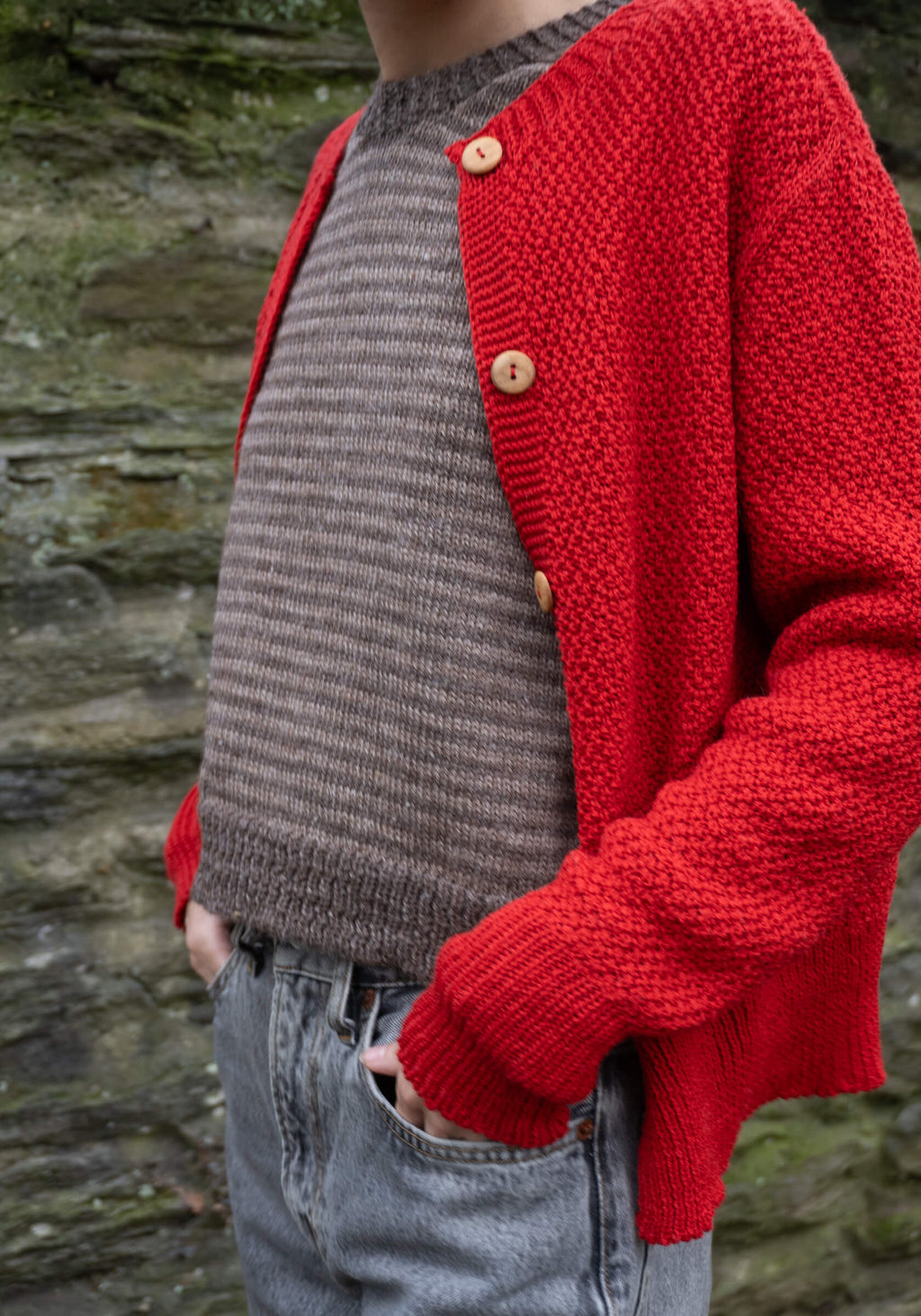 Person wearing a red knitted cardigan over a brown sweater against a stone wall background