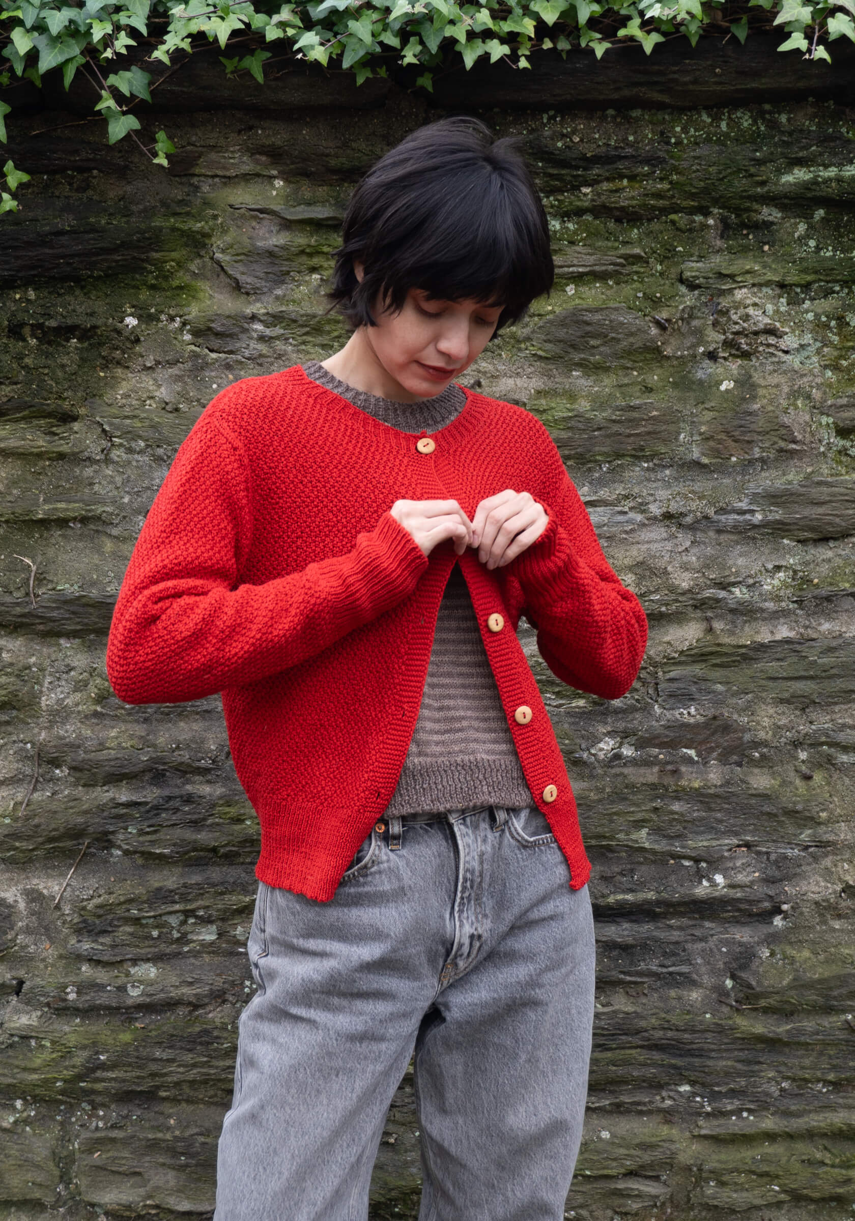 Person wearing a red knitted cardigan over a gray sweater, standing against a stone wall with greenery.
